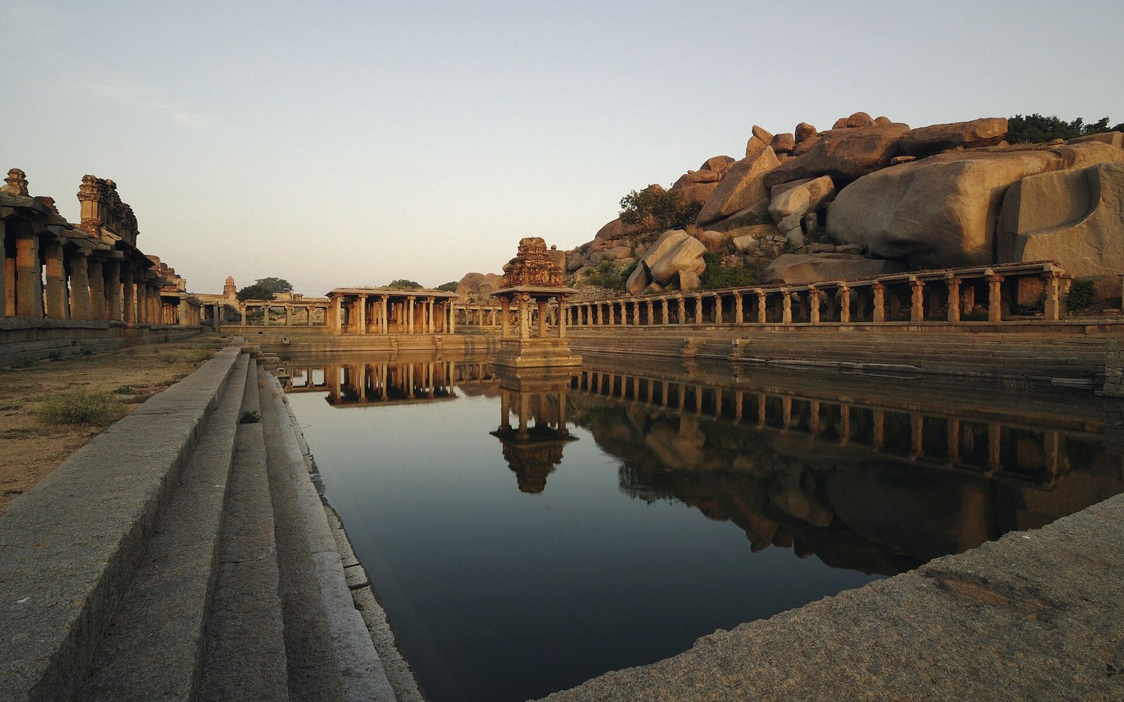Stepped stone tank at Hampi reflects a small pillared pavilion beside boulder-strewn hills